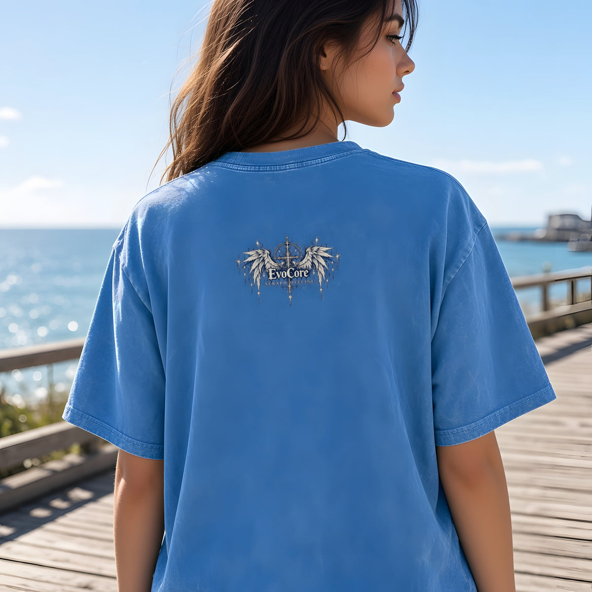 EvoCore Seraph Bluehour Tee in Light Blue, back view worn by a model, seaside boardwalk on a sunny day
