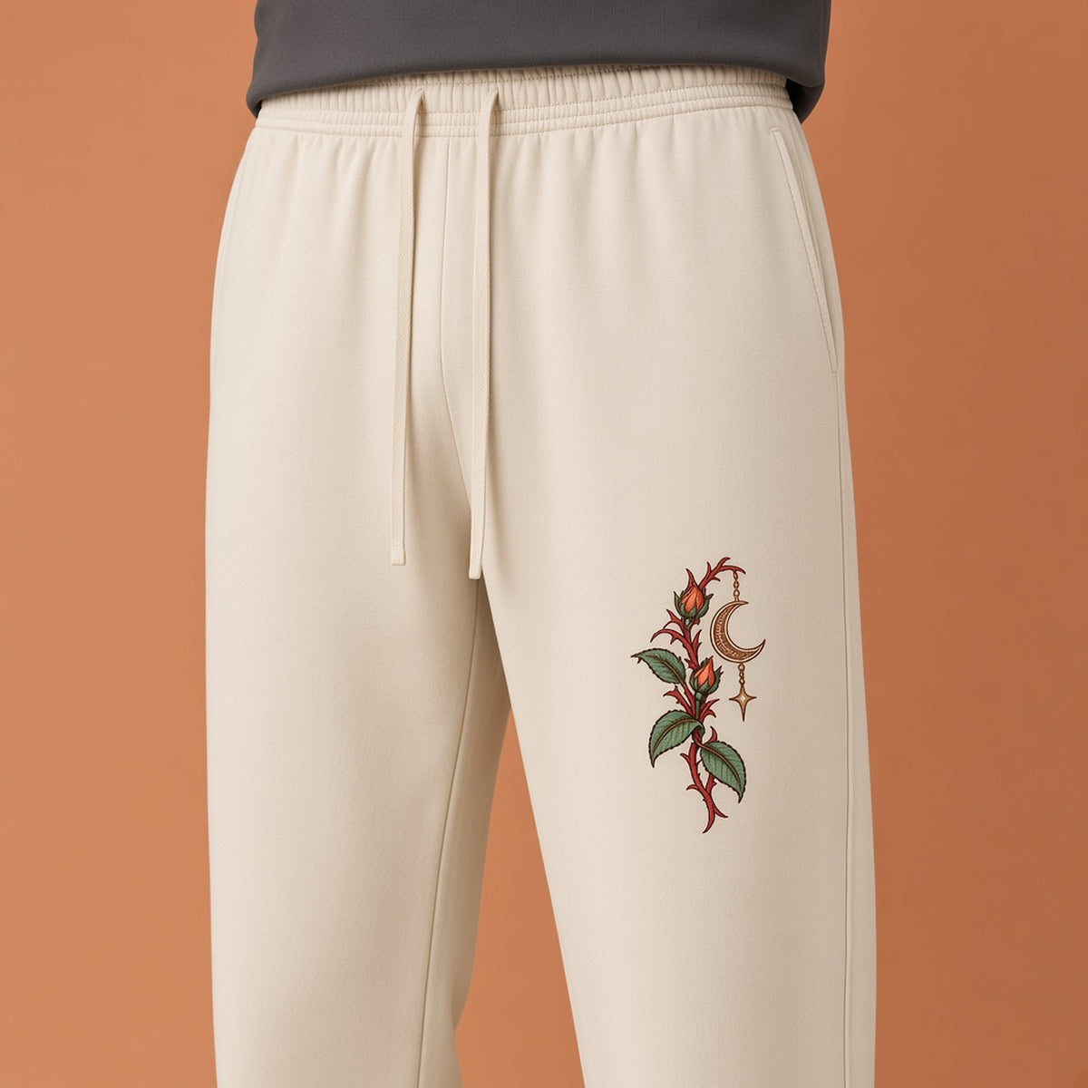 Close-up of the red thorned stem, green leaves and crescent print on EvoCore Thornlight Conservatory Apricot sweatpants against an apricot studio backdrop.